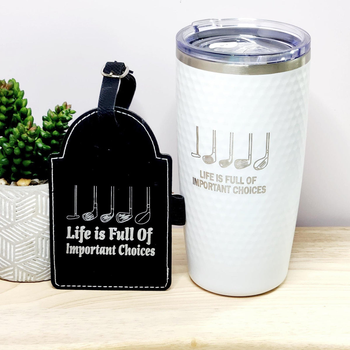 A white stainless steel golf tumbler with text 'Life is Full of Important Choices' and a dimpled texture, accompanied by a black golf tee holder with the same engraving.