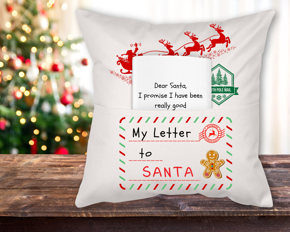A white linen reading pillow with a 'My Letter to Santa' theme, featuring red and green decorative elements and a Ginger Bread Man and the text 'Dear Santa, I promise I have been really good' displayed on it, placed on a wooden surface with a Christmas tree in the background.
