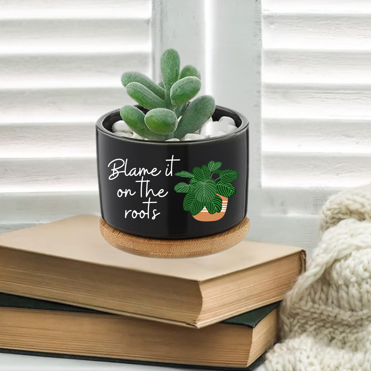 Black ceramic cylinder planter with bamboo saucer reading Blame it on the Roots with illustrated plant graphic, styled on stacked books with a succulent planted inside