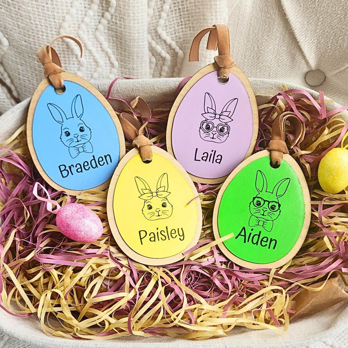 A collection of colorful egg-shaped personalized Easter basket tags with names engraved on them, displayed on a bed of decorative straw.