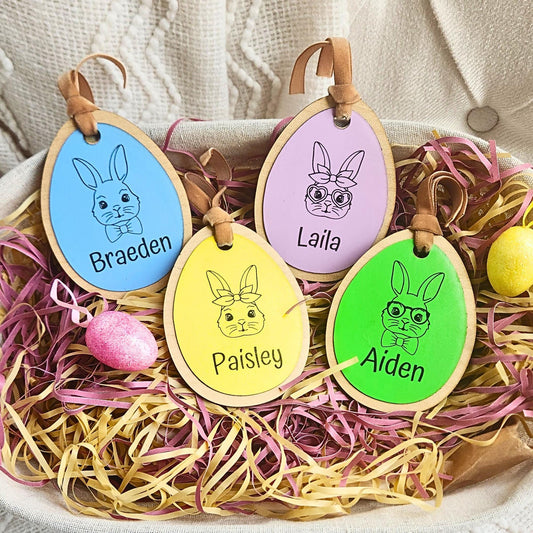 A collection of colorful egg-shaped personalized Easter basket tags with names engraved on them, displayed on a bed of decorative straw.