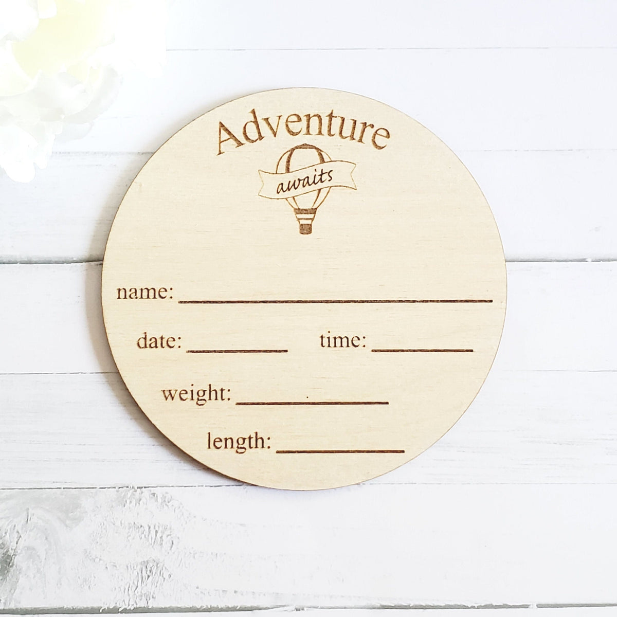 A wooden round disc with 'Adventure Awaits' text and spaces for recording a baby's name, date, time, weight, and length.