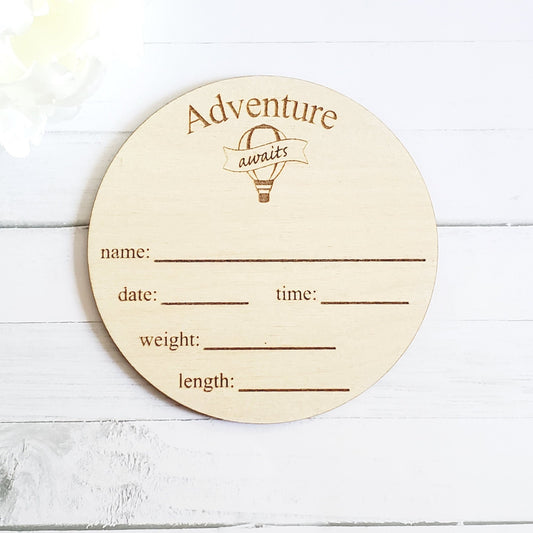 A wooden round disc with 'Adventure Awaits' text and spaces for recording a baby's name, date, time, weight, and length.