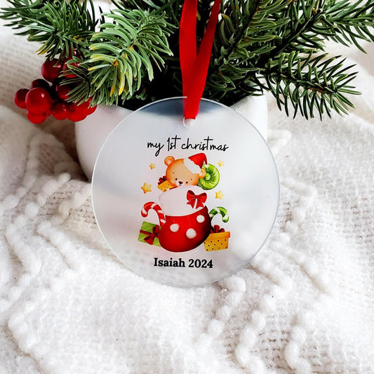A personalized glass ornament with a illustration of a baby in festive attire, with 'my 1st christmas Isaiah 2024' printed on it, attached to a red satin ribbon.