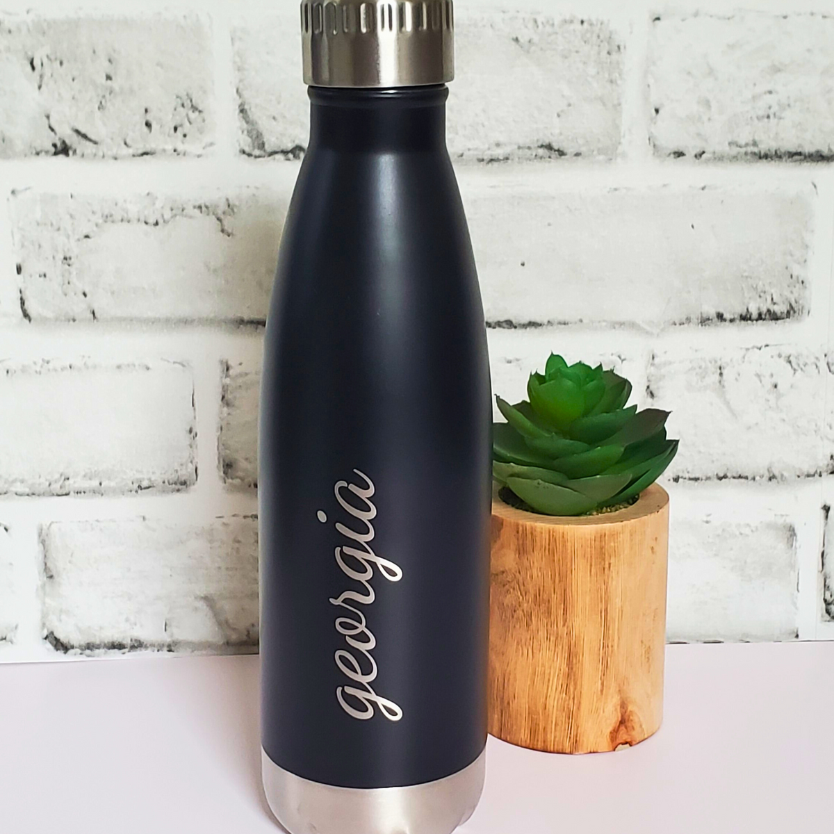 Black water bottle with 'georgia' engraving next to a small green plant on a white brick background