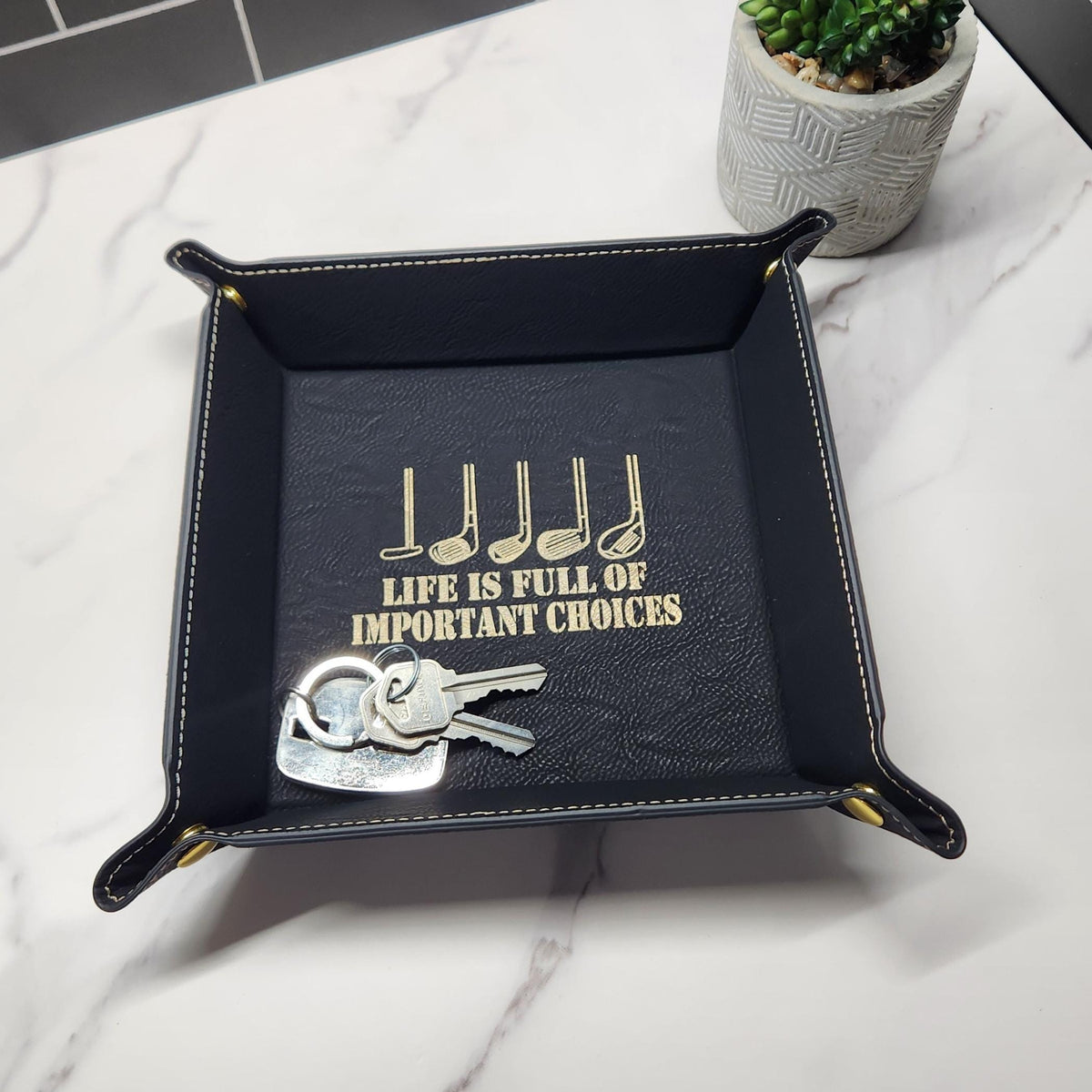 Black vegan leather valet tray holding keys, with gold golf club engraving and the message ‘Life is full of important choices.’