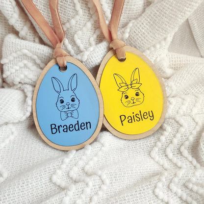 Two personalized Easter basket tags in blue and yellow vegan leather with engraved bunny illustrations and names Braeden and Paisley, tied with ribbon on white knit background.