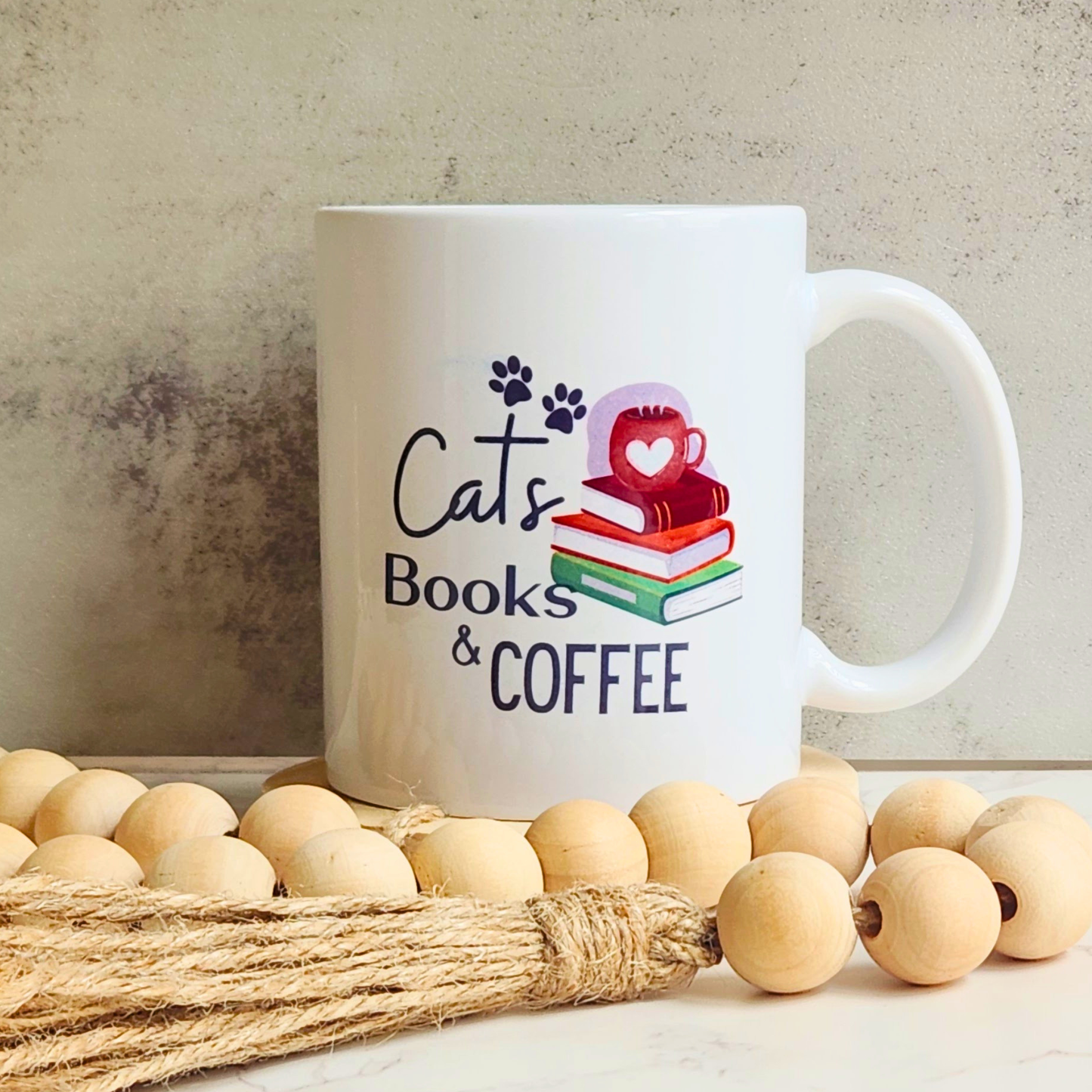 A white ceramic coffee mug with a 'Cats Books & COFFEE' graphic, styled on a tabletop with wooden beads and a jute rope against a textured concrete wall.