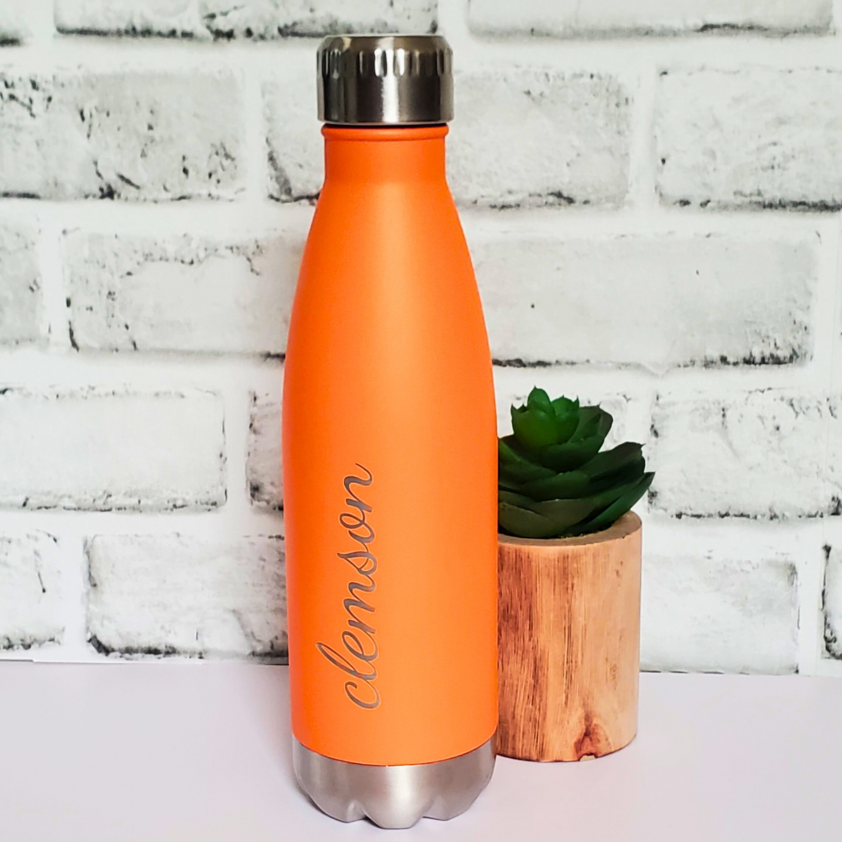 Orange water bottle with 'Clemson' engraving next to a small plant against a white brick wall.