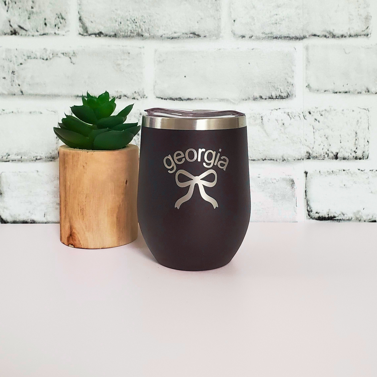 Black tumbler with 'georgia' and a bow design on a white surface with a white brick wall background.