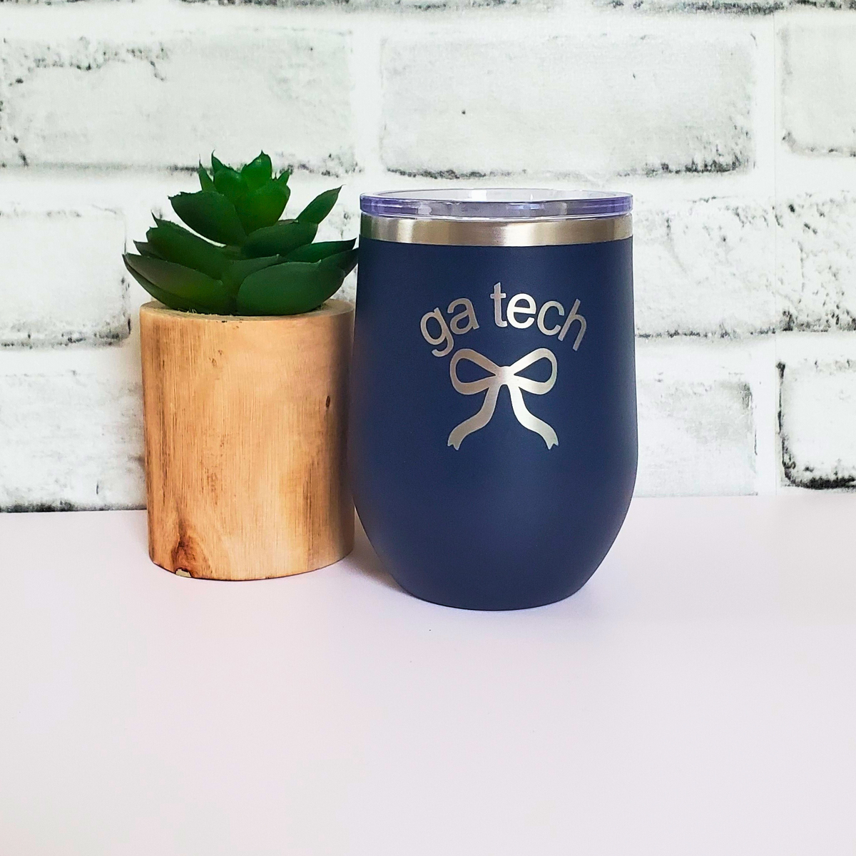 Navy blue tumbler with 'ga tech' and a bow design next to a wooden planter with a plant on a white brick background