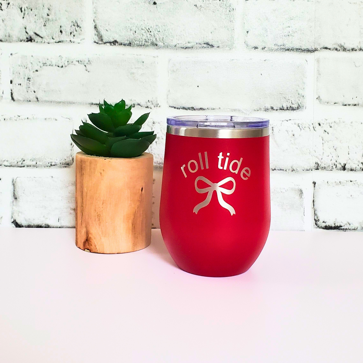 Red wine tumbler with 'roll tide' design next to a small potted plant on a white surface with a white brick wall background.