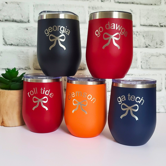 Five insulated wine tumblers in black, red, orange, and blue with text and bow designs on a white brick background.