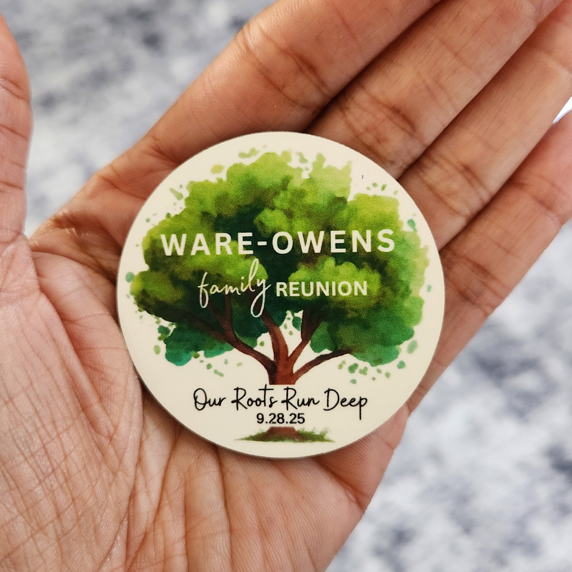 Hand holding a round family reunion magnet with 'Ware-Owens' family name and "Our Roots Run Deep" and event date with green tree in the background. 