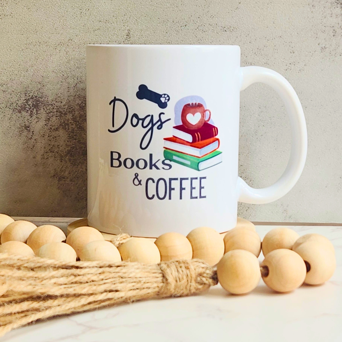 A white ceramic mug with a 'Dogs Books & COFFEE' graphic, styled on a marble surface with wooden beads and a jute rope against a textured concrete wall.