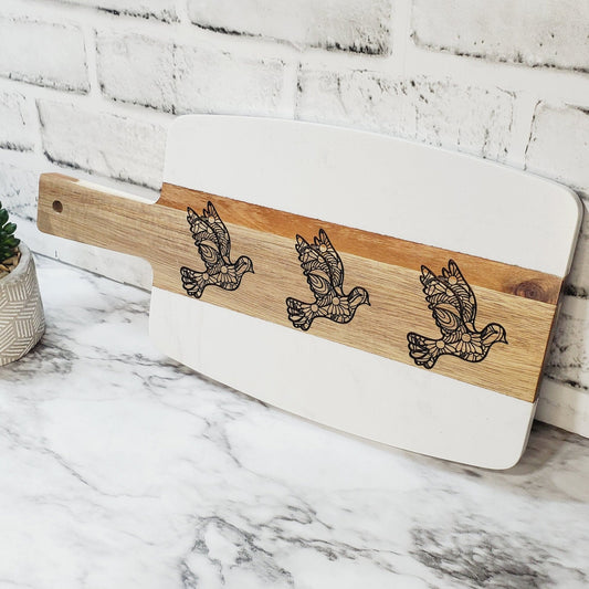 A marble and acacia wood charcuterie board with engraved doves on the wood surface, displayed on a white surface with a brick wall in the background.