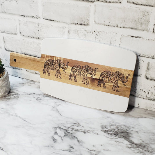 A marble charcuterie board with acacia wood handles and elephant engravings, placed against a white brick wall on a marble surface.