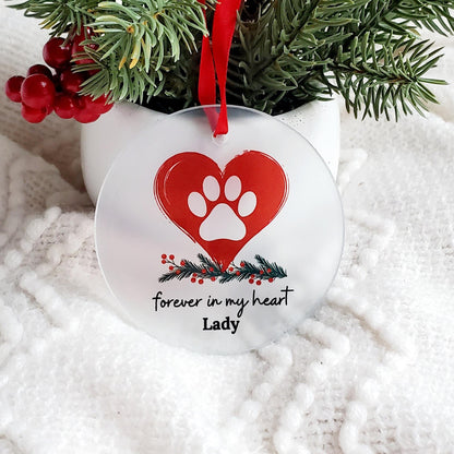 A glass circular pet ornament with a red heart and a white paw print, personalized with the name 'Lady'. The ornament is hanging from a red satin ribbon, with decorations of green foliage and red berries in the background.