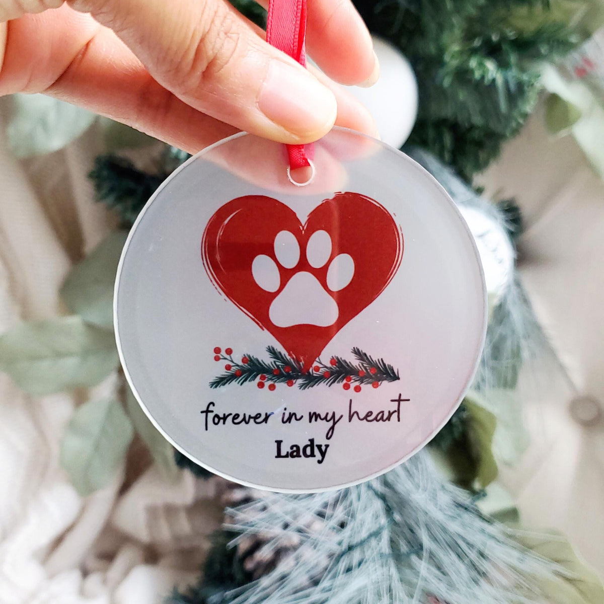 A glass circular pet ornament with a red heart and a white paw print, personalized with the name 'Lady'. The ornament is hanging from a red satin ribbon, with decorations of green foliage and red berries in the background.