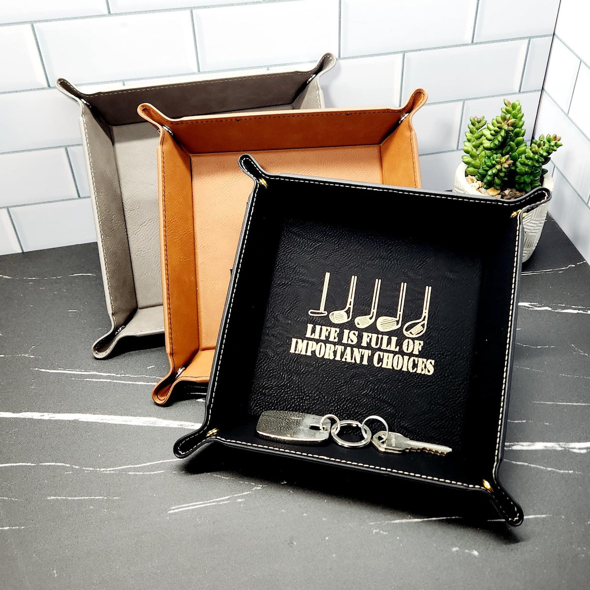 Black, tan, and grey vegan leather valet trays, with keys and a gold golf-themed engraving visible in the front black tray.