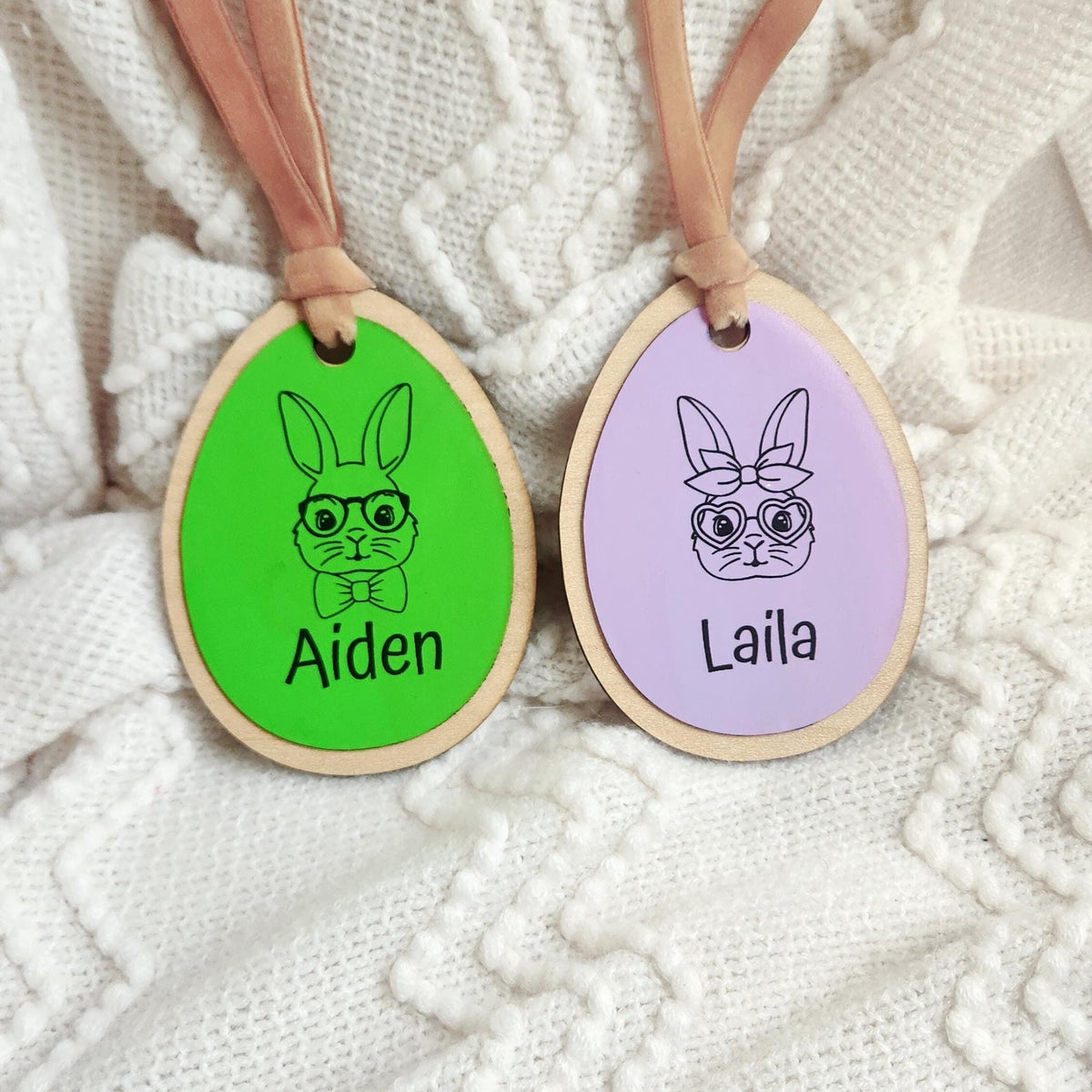 Close-up of two personalized Easter basket tags — green with engraved boy bunny for Aiden and lilac with girl bunny for Laila — on white textured background.