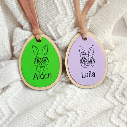 Close-up of two personalized Easter basket tags — green with engraved boy bunny for Aiden and lilac with girl bunny for Laila — on white textured background.