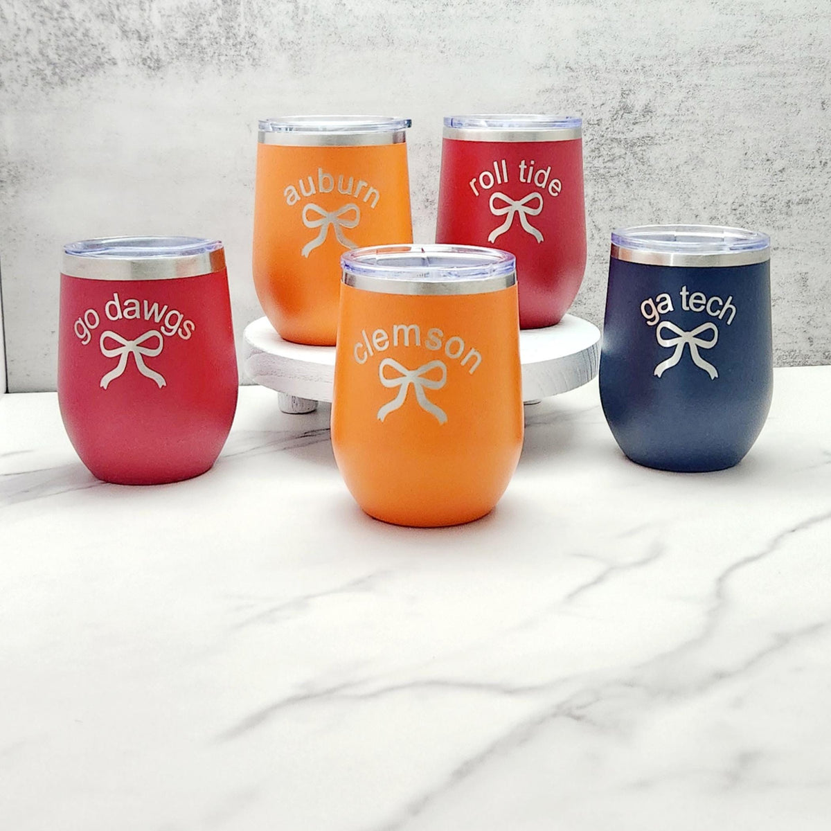 A group of wine tumblers engraved with a silver bow and printed red "go dawgs", orange "clemson", orange "auburn" , blue "ga tech", and red "roll tide".
