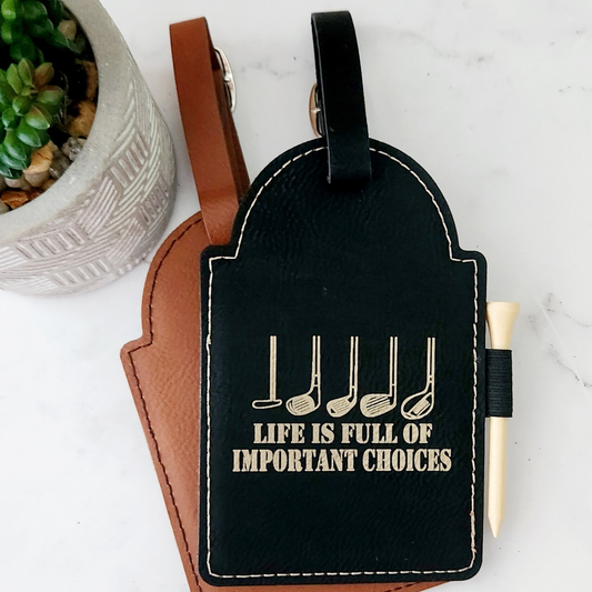 Black vegan leather golf bag tag with gold golf clubs and ‘Life is full of important choices’ engraving, layered over a tan tag.