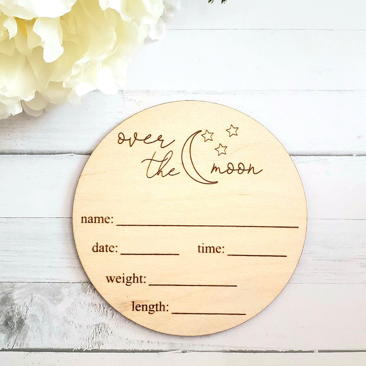 A wooden round disc with 'over the moon' text and stars, intended for capturing baby milestones such as name, date, time, weight, and length.