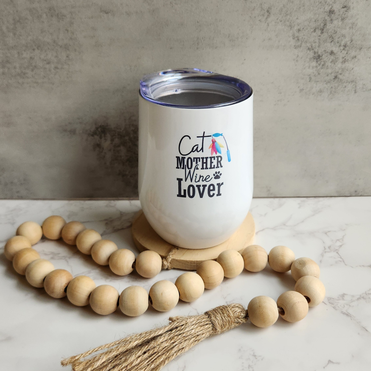 White 12 oz Cat Mother Wine Lover insulated tumbler with colorful feather wand graphic, styled on a marble surface with a wooden bead garland