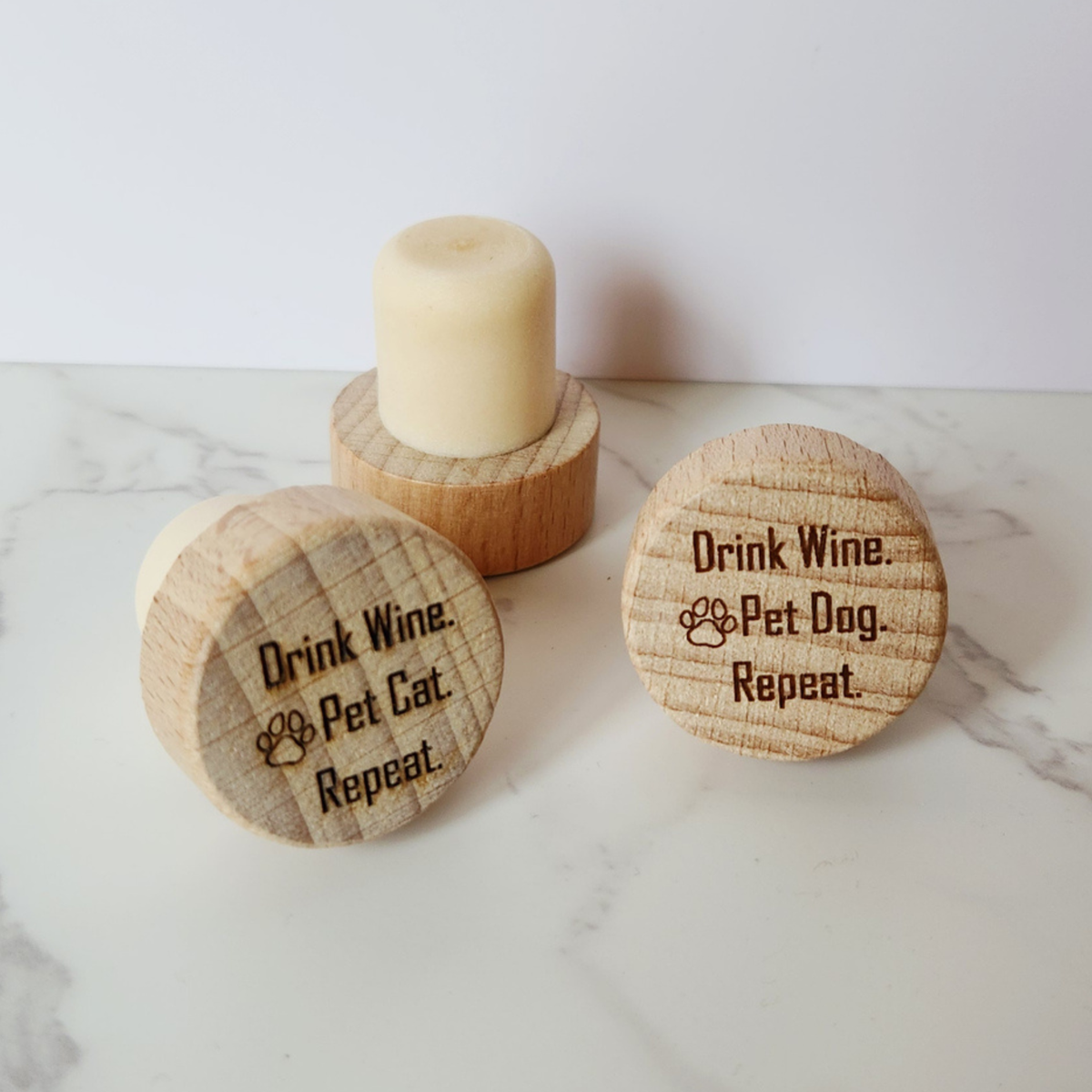 Three round wooden wine cork stoppers engraved text reading 'Drink Wine & Pet Cat. Repeat.' and 'Drink Wine & Pet Dog. Repeat.'