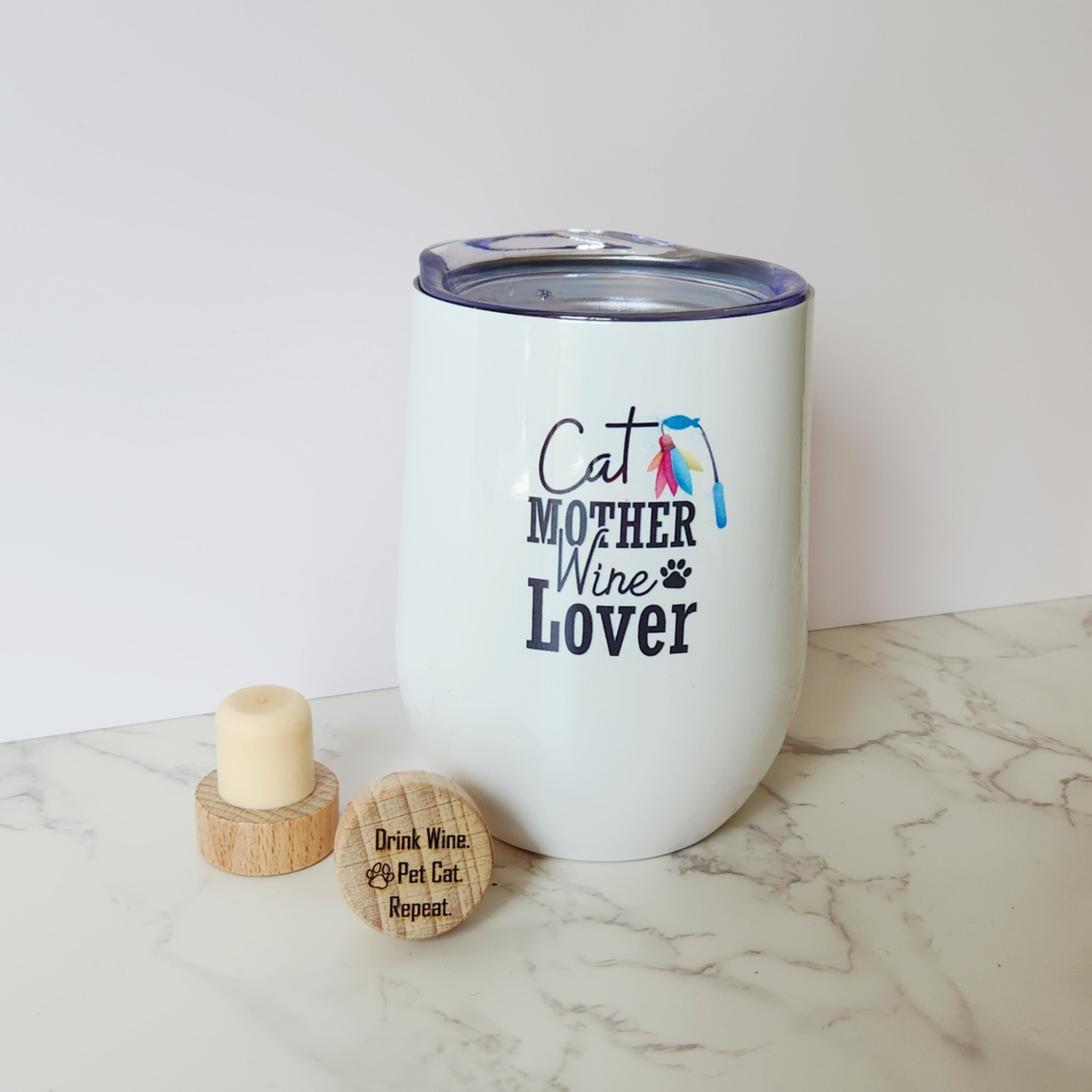 A white insulated stemless wine tumbler with a clear lid, printed with the text 'Cat MOTHER Wine Lover' and a small colorful cat graphic, shown on a marble surface with two wooden wine stoppers beside it.