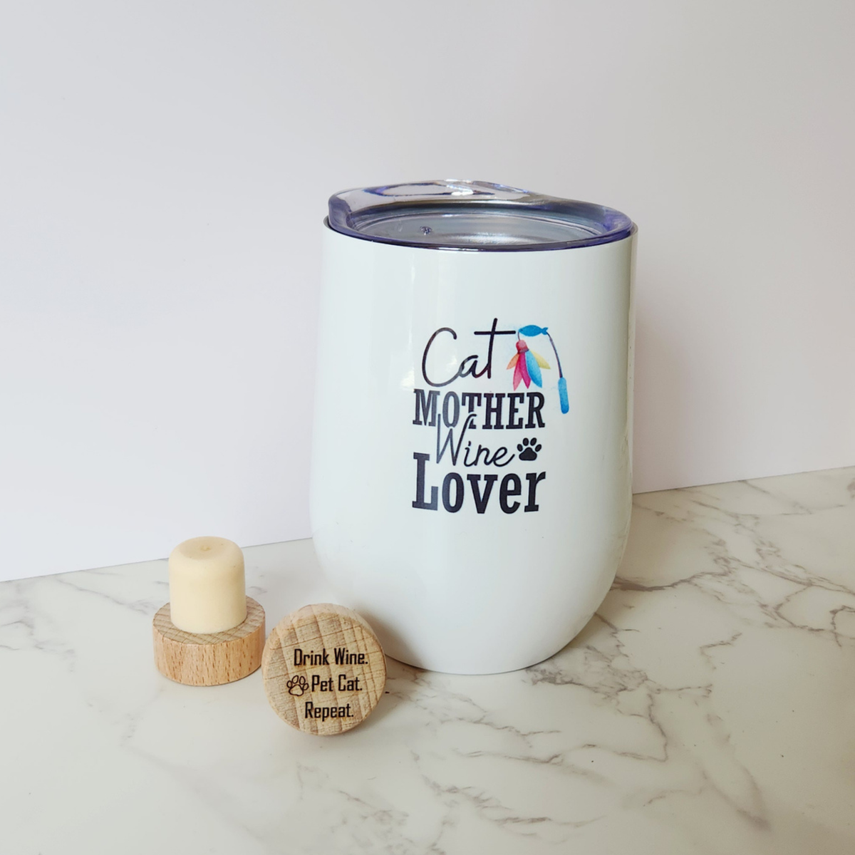 A white insulated stemless wine tumbler with a clear lid, printed with the text 'Cat MOTHER Wine Lover' and a small colorful cat graphic, shown on a marble surface with two wooden wine stoppers beside it.