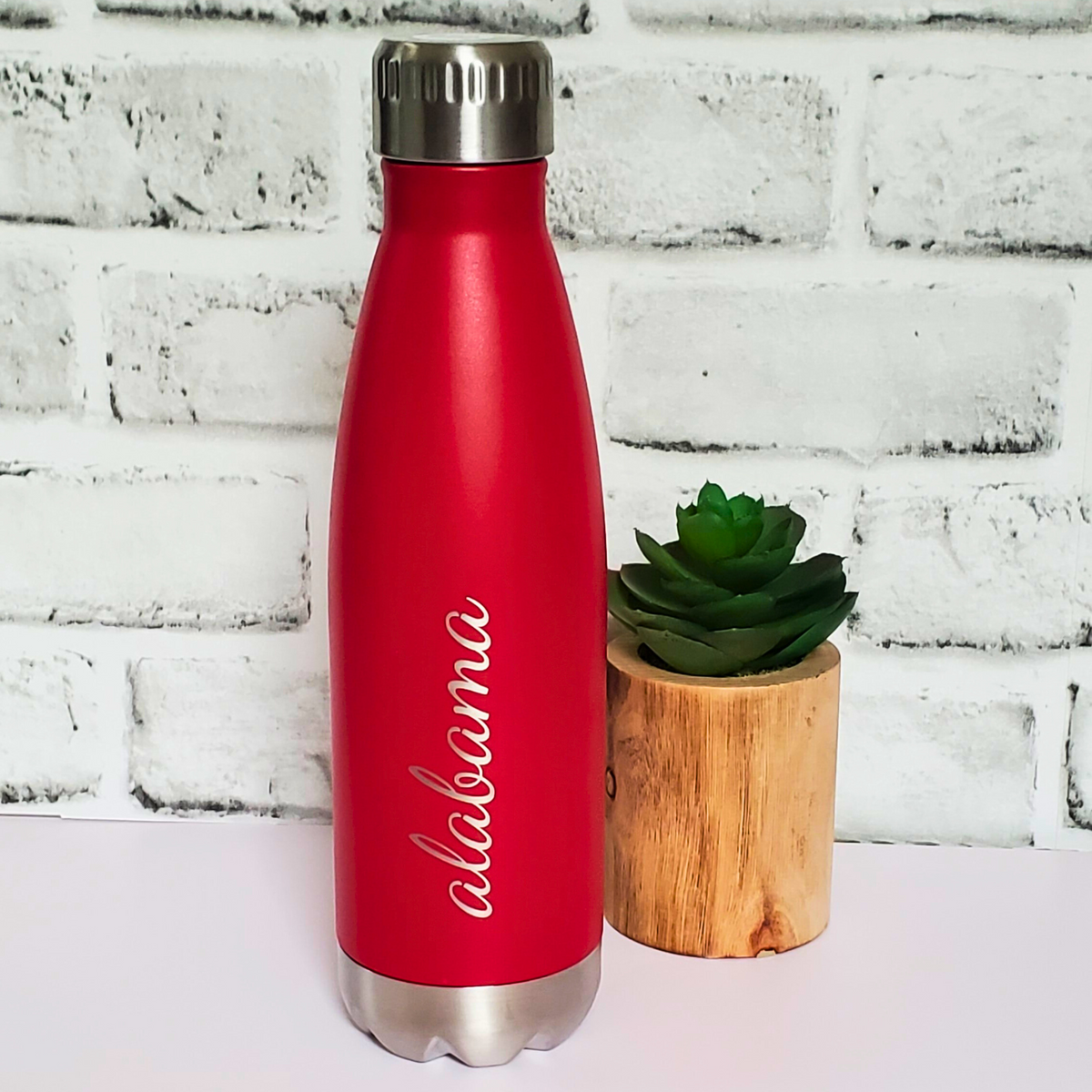 Red water bottle with 'Alabama' text next to a small potted plant against a white brick wall.