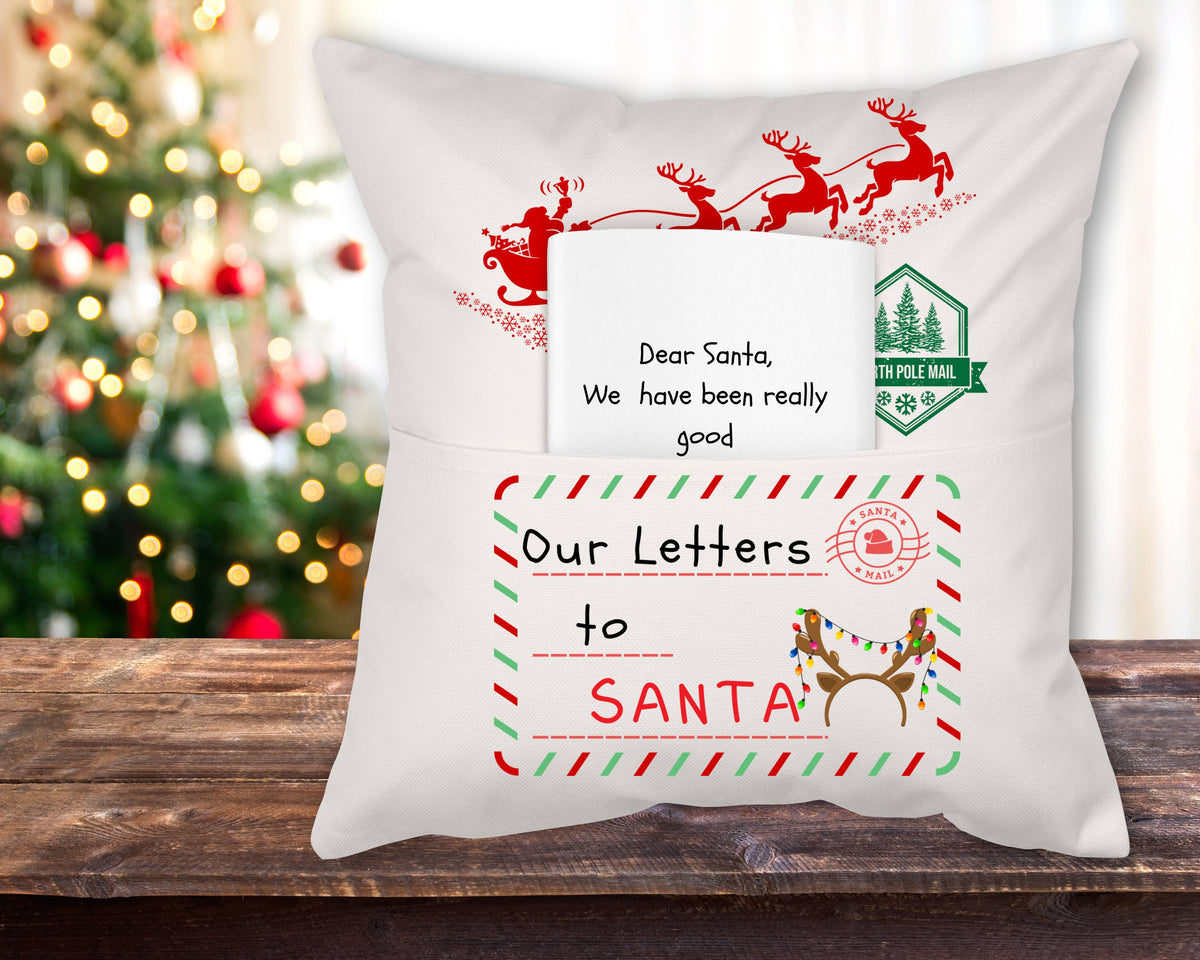 A white linen reading pillow with a 'Our Letter to Santa' theme, featuring red and green decorative elements and the text 'Dear Santa, I promise I have been really good' displayed on it, placed on a wooden surface with a Christmas tree in the background.