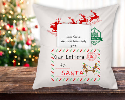 A white linen reading pillow with a 'Our Letter to Santa' theme, featuring red and green decorative elements and the text 'Dear Santa, I promise I have been really good' displayed on it, placed on a wooden surface with a Christmas tree in the background.