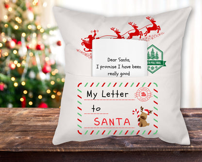 A white linen reading pillow with a 'Letter to Santa' theme, featuring red and green decorative elements and the text 'Dear Santa, I promise I have been really good' displayed on it, placed on a wooden surface with a Christmas tree in the background.