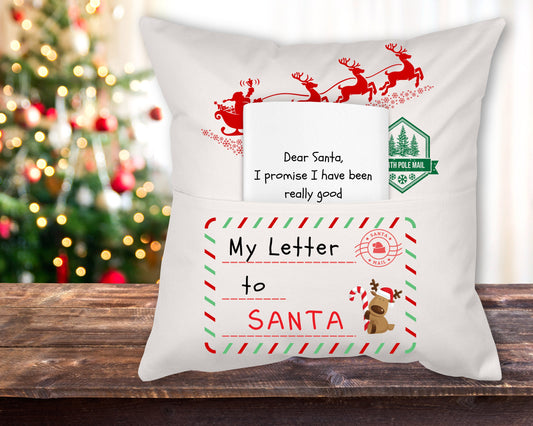 A white linen reading pillow with a 'Letter to Santa' theme, featuring red and green decorative elements and the text 'Dear Santa, I promise I have been really good' displayed on it, placed on a wooden surface with a Christmas tree in the background.