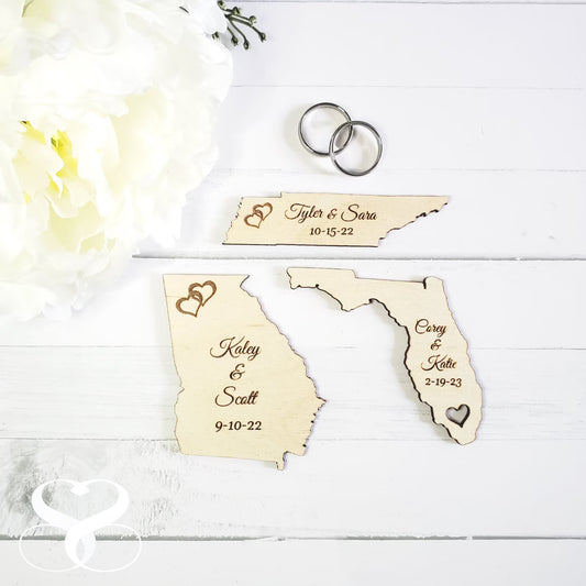 Customized wooden state-shaped wedding favor magnets with engraved hearts and names, displayed on a white surface alongside wedding rings and flowers.