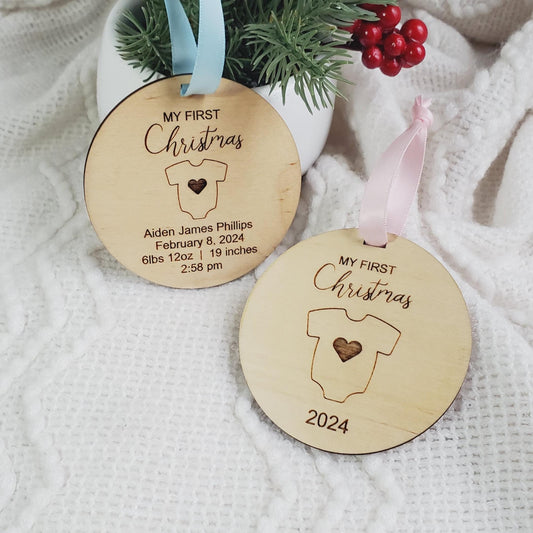 Two wooden Christmas ornaments with 'MY FIRST Christmas' engraved on them, one with a blue ribbon and the other with a pink ribbon, displayed on a white fabric surface.