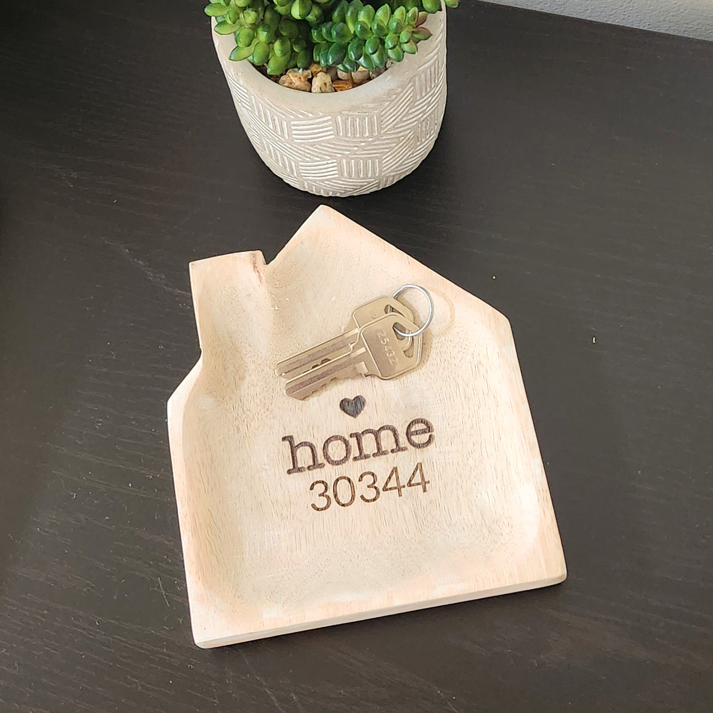 Light colored wooden tray in the shape of a house. Engraved with heart above the word home.  And a zipcode 30344 underneath the word home.  This tray holds 2 keys on a keyring.