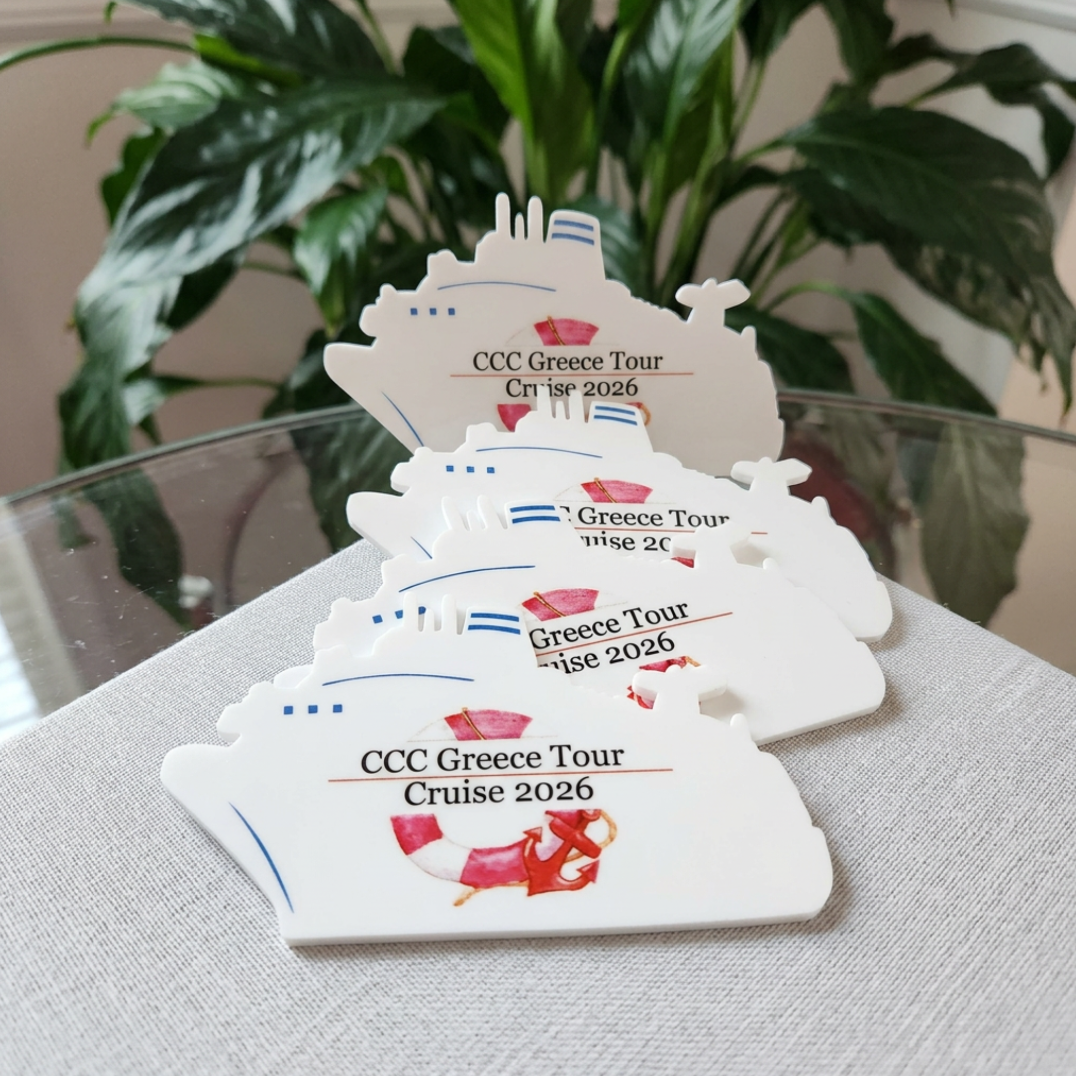 Four acrylic cutouts shaped like cruise ships are stacked on a light gray fabric surface, with a background of green houseplants and a glass table. Each cutout has printed text reading 'CCC Greece Tour Cruise 2026' with small red and blue graphic accents.